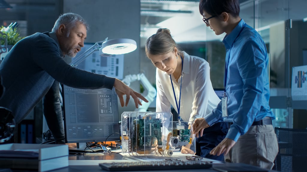 Team of Computer Engineers Lean on the Desk and Choose Printed Circuit Boards to Work with, Computer Shows Programming in Progress. In The Background Technologically Advanced Scientific Research Center.